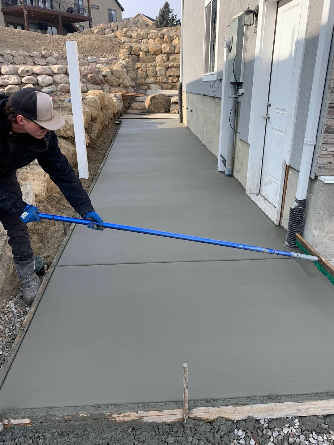 Worker smoothing freshly poured concrete sidewalk near a house foundation.