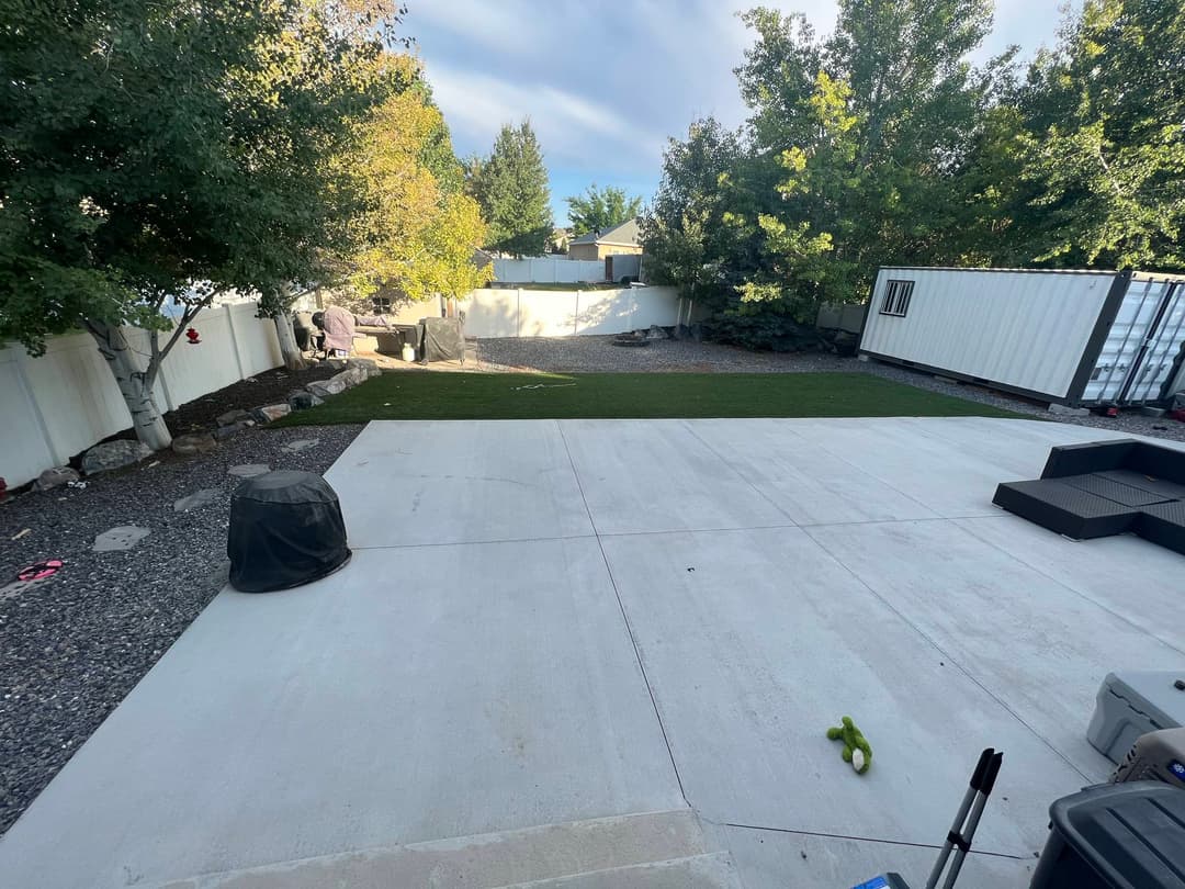 Backyard with new turf, concrete patio, and storage container, surrounded by trees and gravel.
