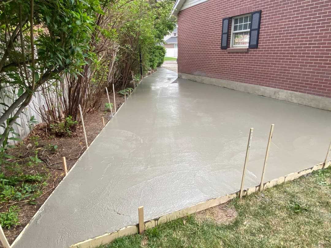 Freshly poured concrete walkway beside a brick house, surrounded by greenery and wooden stakes.