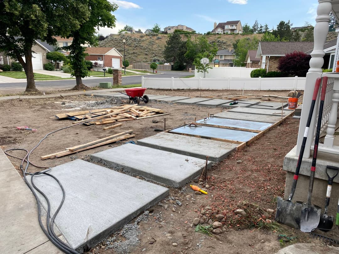 Concrete sidewalk installation in residential area with construction materials and tools.