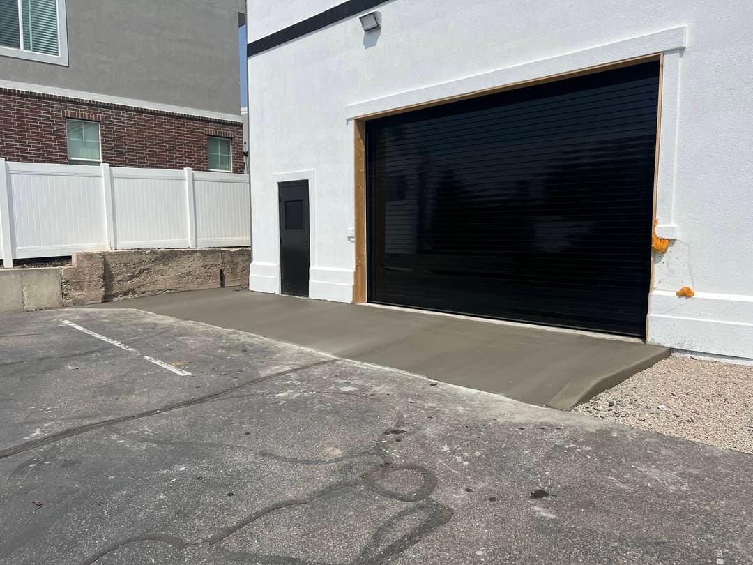 Freshly poured concrete slab in front of a garage door, with a white building and gray fence.