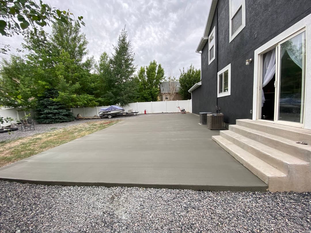 Freshly poured concrete patio with steps beside a gray house and landscaped yard.