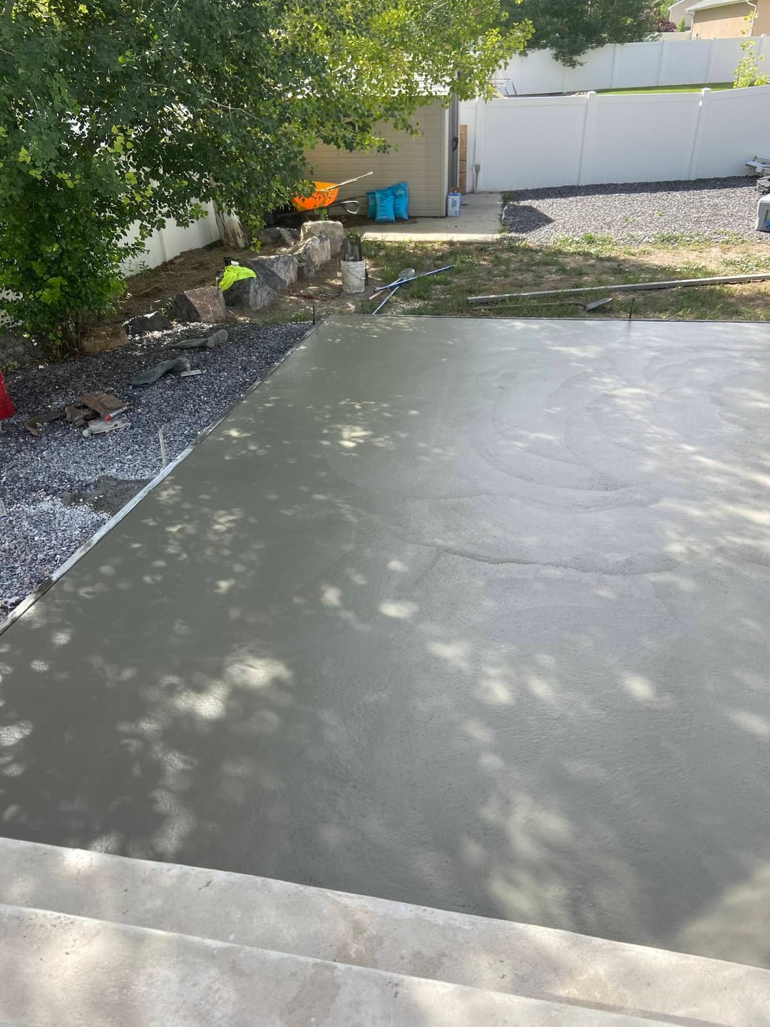 Freshly poured concrete slab in a yard under a tree, surrounded by gravel and tools.