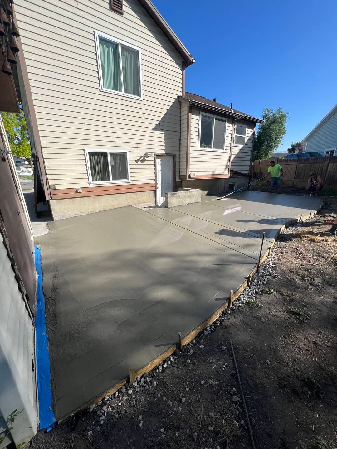 Freshly poured concrete patio installation beside a two-story house with construction workers nearby.