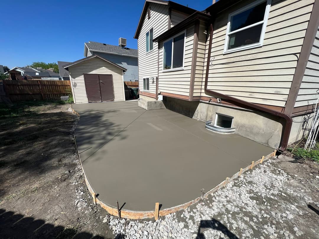 Freshly poured concrete patio extension beside a house and storage shed in a residential backyard.